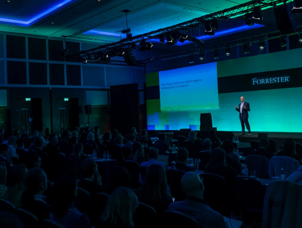 Forrester conference speaker presenting on stage to a large audience at a B2B business event with blue and green event lighting