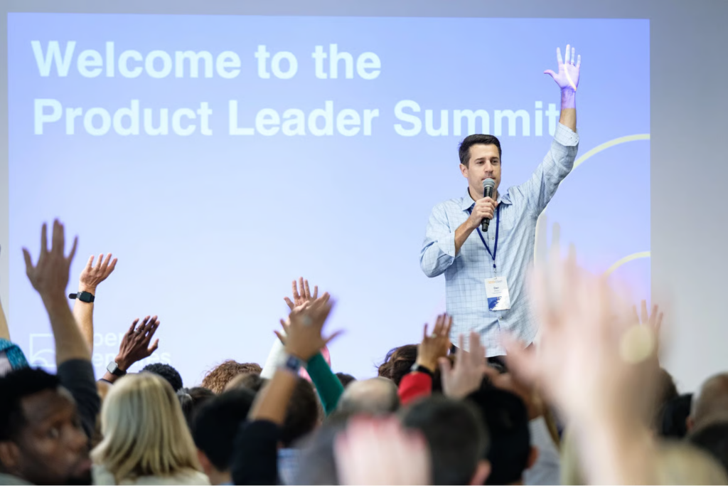 Ponente en el escenario con micrófono y mano levantada dirige la palabra al público en el Product Leader Summit.